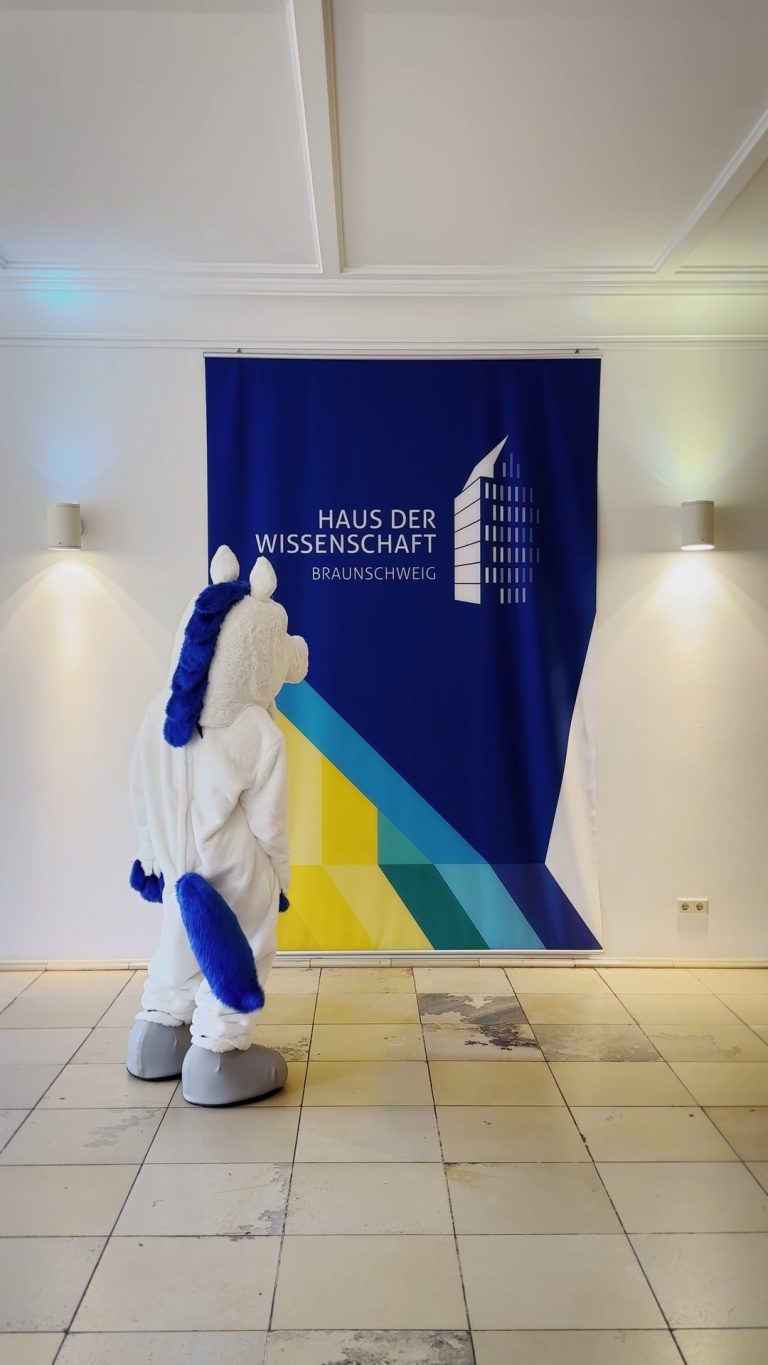 Maskottchen der Öffentlichen Versicherung Braunschweig Öfi vor einem Banner vom Haus der Wissenschaft